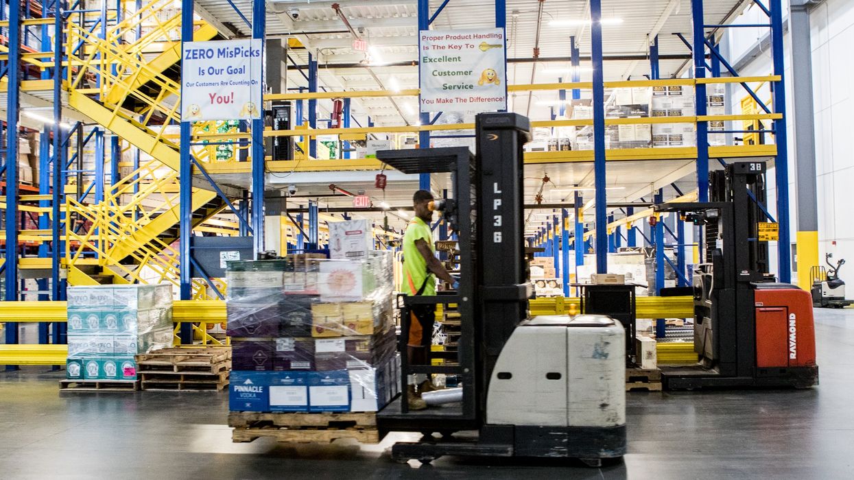 A man drives a stand-up lift truck in a warehouse. Another lift truck, pallets, and racks are nearby.